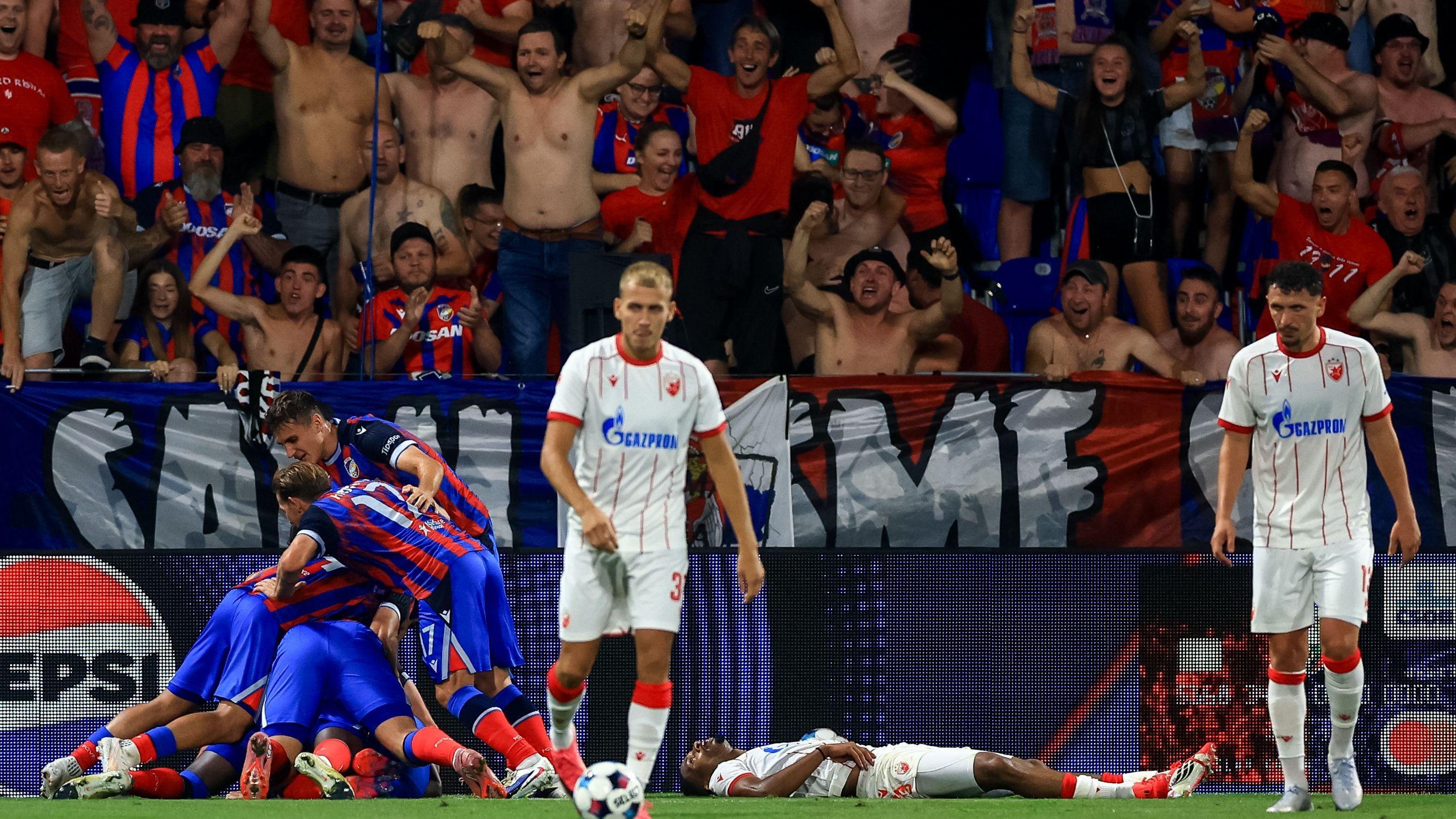 fudbaleri na terenu, sa leve strane u plavo crvenim dresovima, desno dva fubalera u belim dresovima sa crvenim uspravnim linijama okrenuta ka kameri, jedan šutira loptu, između njih leži fudbaler na terenu