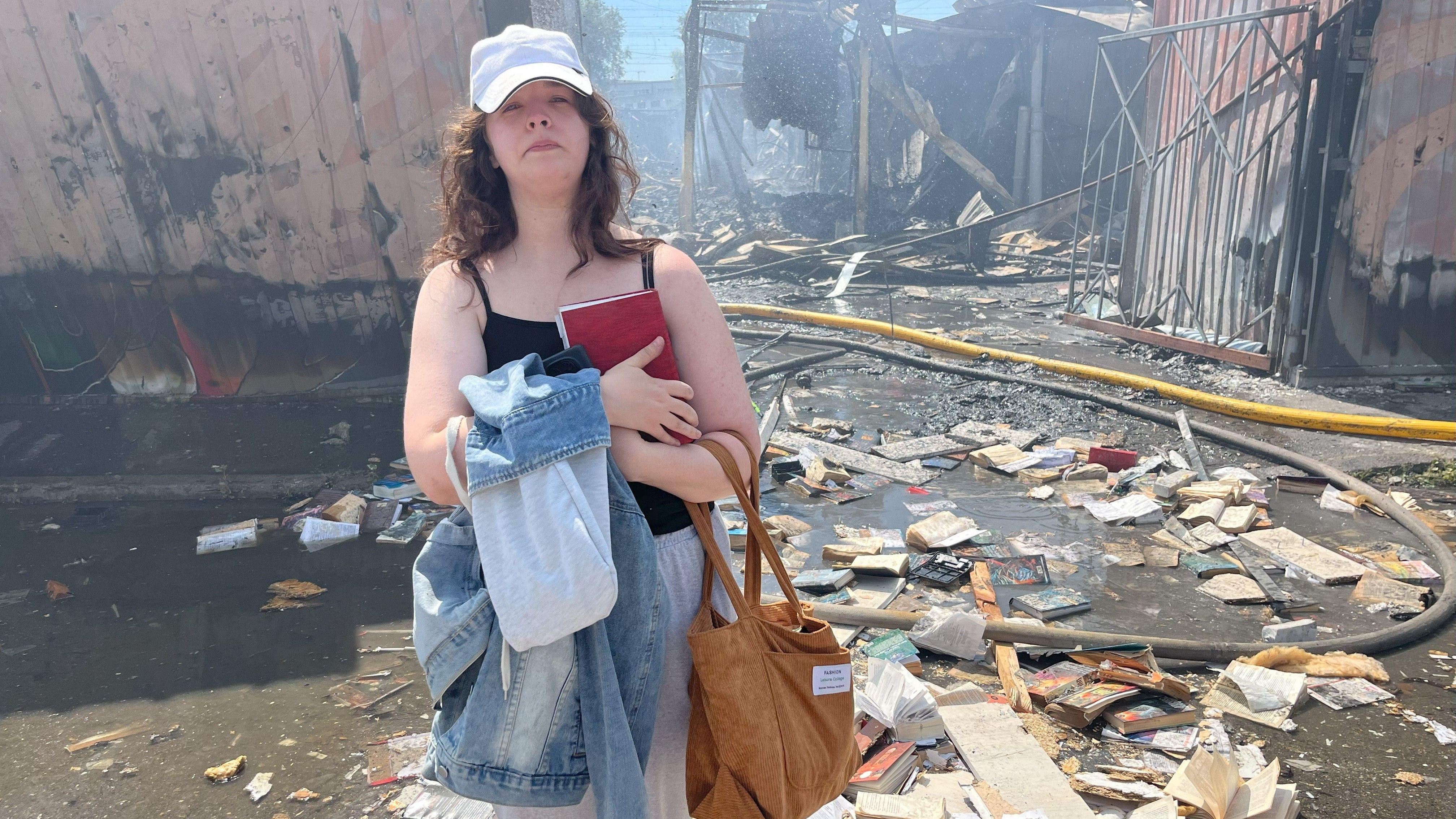 Mlada žena čvrsto drži knjigu na grudima i izgleda tužno. Iza nje je bombardovana pijačna građevina. Uništene knjige razbacane su po zemlji oko nje.