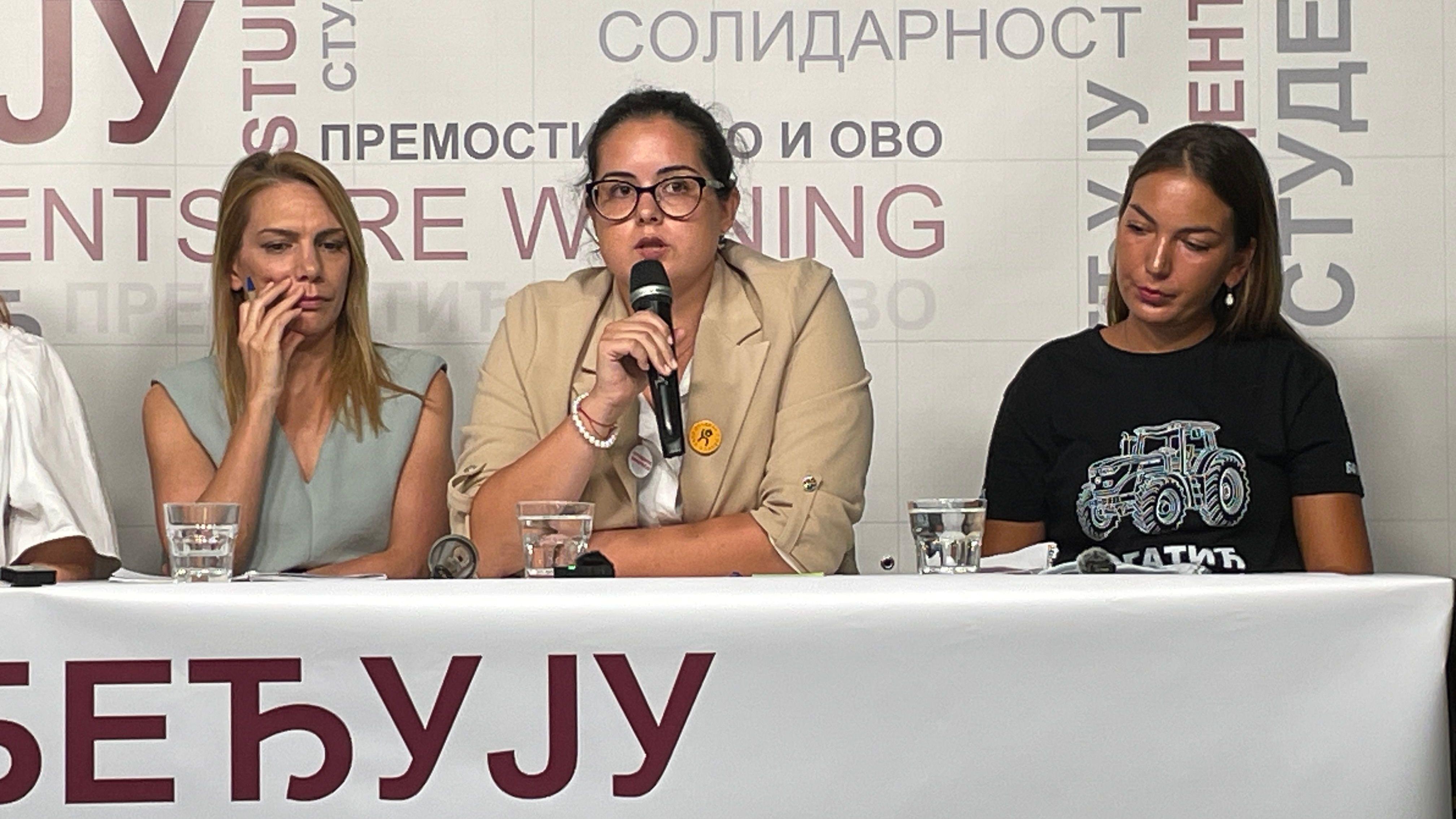 Devojka na konferenciji govori u mikrofon, ima vezanu tamnu kosu, naočale tamne boje i obučena je u svetlu garderobu