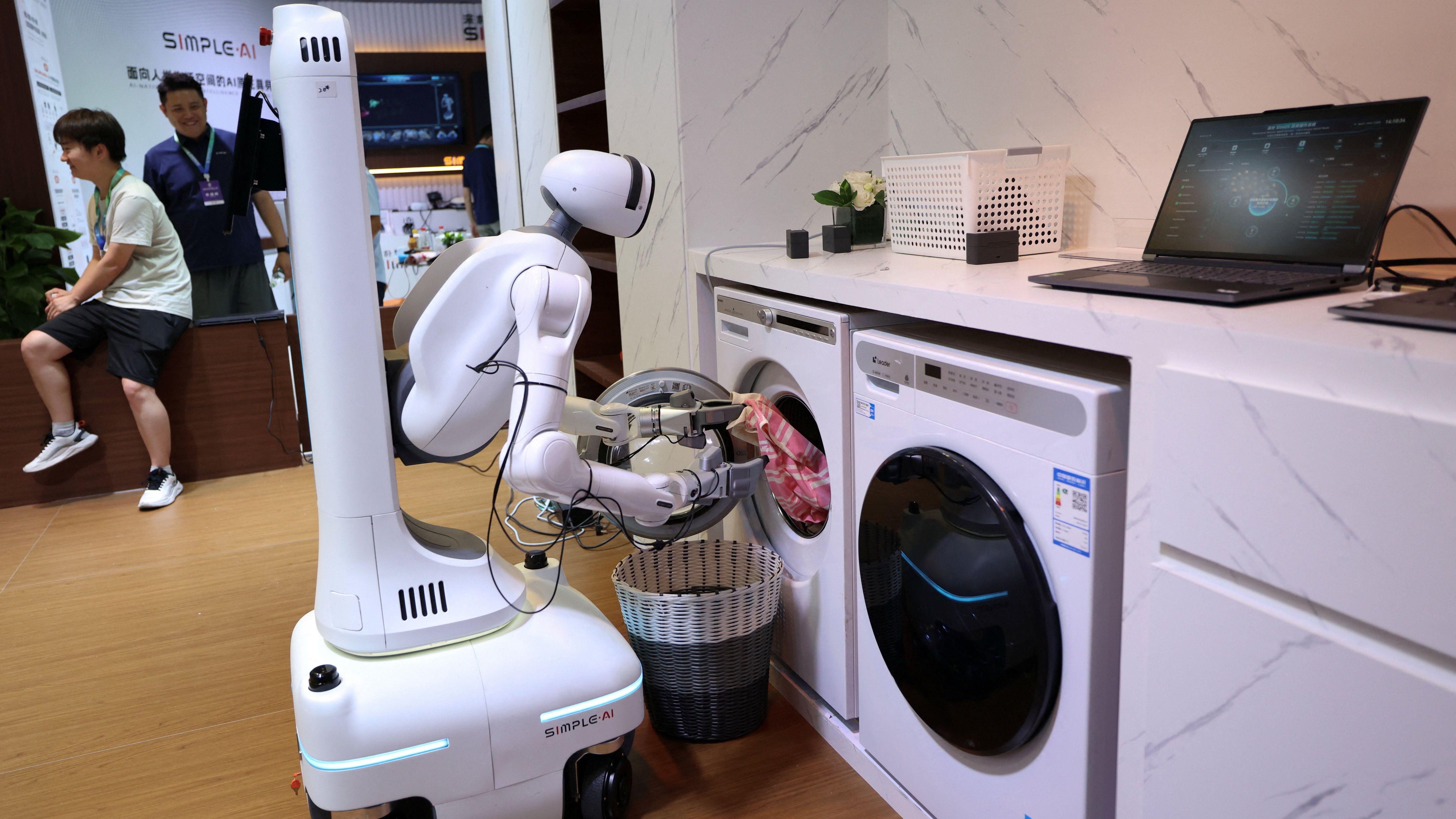 Beli robot sa humanoidnim torzom istovaruje mašinu za veš u maketu kuhinje na izložbi, dok posetilac sa priveskom posmatra