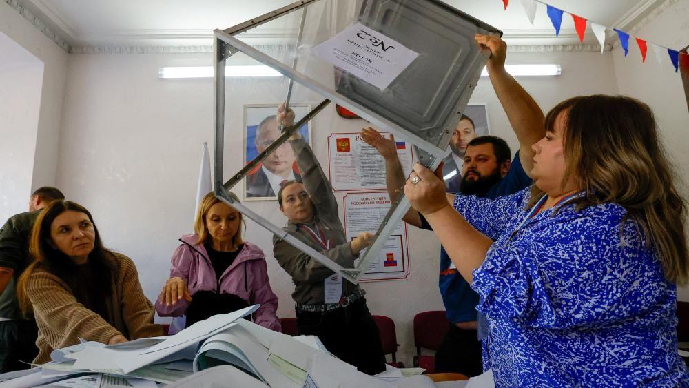 Članovi lokalne izborne komisije prazne glasačke kutije posle izbora, dok je iza njih velika slika Vladimira Putina