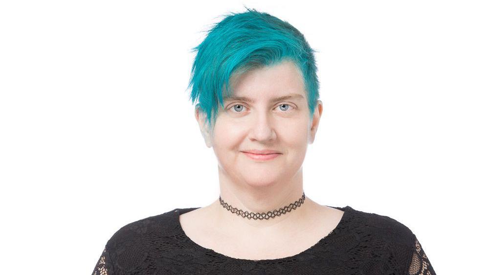 Žena kratke tirkizno plave kose gleda u kameru i smeši se, ima plave oči i tamnu majicu