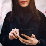 Žena duge tamne kose, u crnom kaputu, gleda u telefon. Iza nje su nacrtani spermatozoida i fotografija ruke koja drži praznu posudicu za uzorke sa crvenim poklopcem