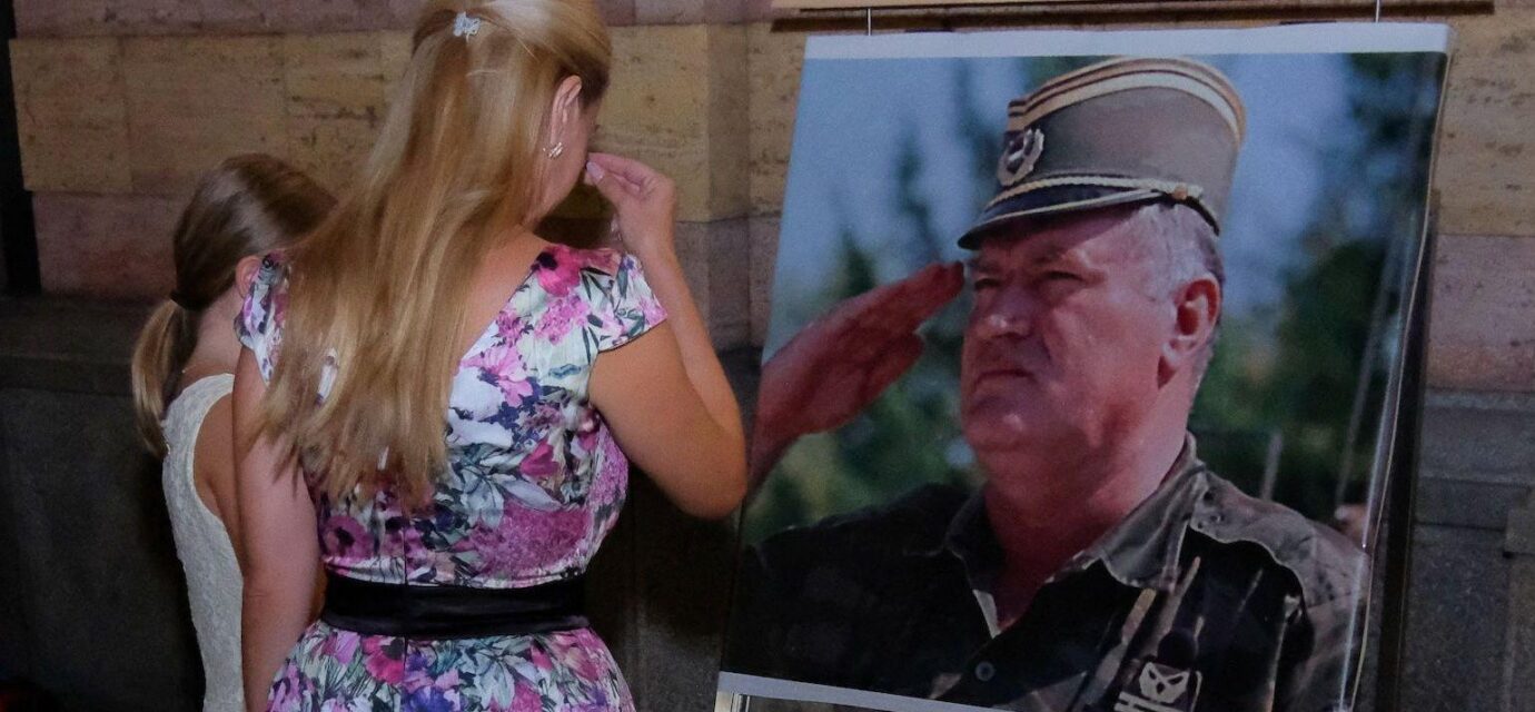 Žena u šarenoj haljini i devojčica u beloj stoje ispred velike fotografije Ratka Mladića u vojnoj uniformi i krste se