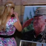 Žena u šarenoj haljini i devojčica u beloj stoje ispred velike fotografije Ratka Mladića u vojnoj uniformi i krste se