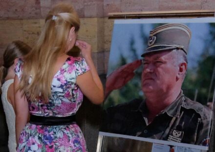 Žena u šarenoj haljini i devojčica u beloj stoje ispred velike fotografije Ratka Mladića u vojnoj uniformi i krste se