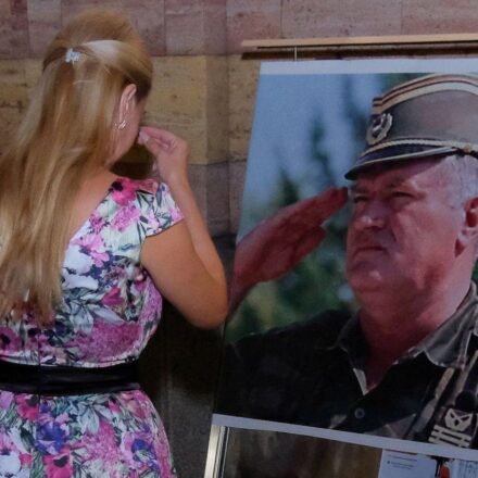 Žena u šarenoj haljini i devojčica u beloj stoje ispred velike fotografije Ratka Mladića u vojnoj uniformi i krste se