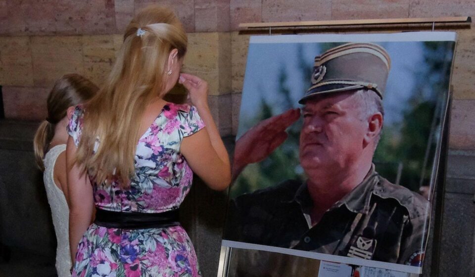 Žena u šarenoj haljini i devojčica u beloj stoje ispred velike fotografije Ratka Mladića u vojnoj uniformi i krste se