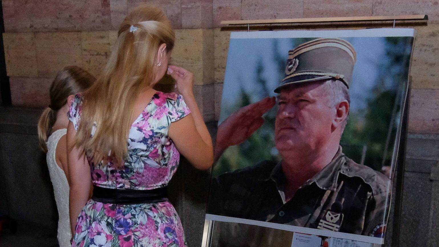 Žena u šarenoj haljini i devojčica u beloj stoje ispred velike fotografije Ratka Mladića u vojnoj uniformi i krste se