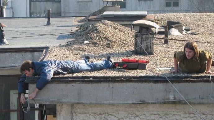 Dva muškarca na krovu zgrade. Jedan, obučen u plavo, leži na krovu i gleda preko ivice dok spušta kablove niz stranu zgrade. Desno od njega nalazi se drugi muškarac duge kose, obučen u zeleno, koji se podigao oslanjajući se na ruke i takođe gleda preko ivice zgrade.