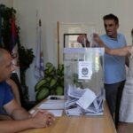 Muškarac za stolom posmatra dvoje koji ubacuju glasački listić u kutiju