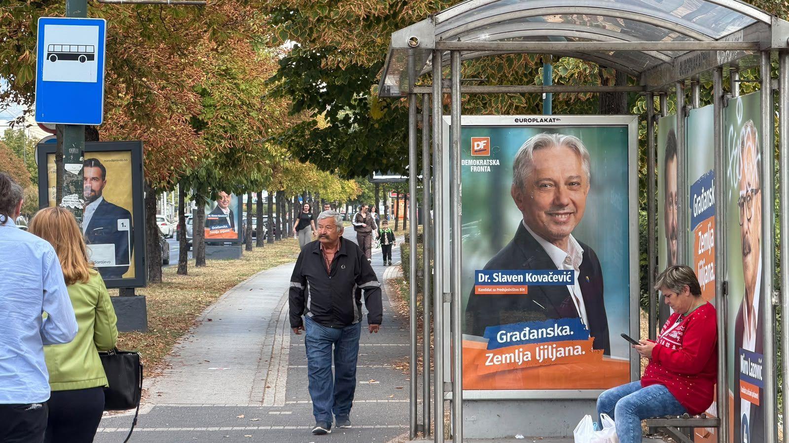 Muškarac hoda ulicom pored gradskog stajališta za prevoz, žena sedi na klupi u crvenom džemperu