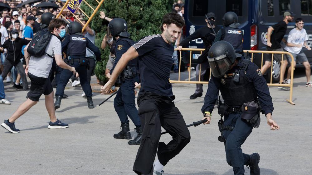 Policajac iz jedinice za razbijanje demonstracija drži palicu blizu zadnje strane nogu jednog demonstranta. U pozadini se vide mase demonstranata i nekoliko policajaca u uniformama i sa šlemovima. Takođe se u pozadini može videti osoba koja podiže deo žute ograde.