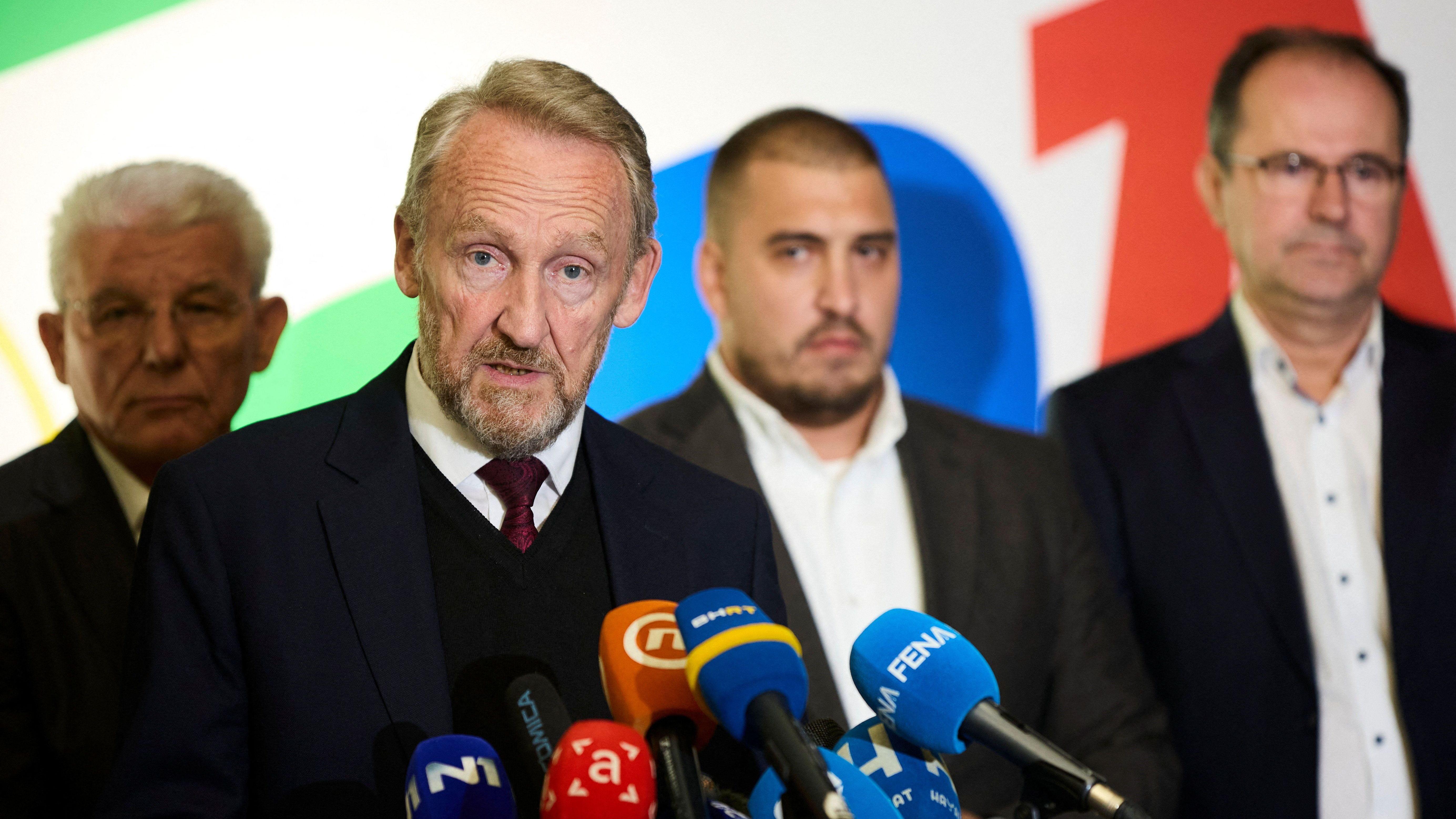 Bakir Izetbegović na konferenciji ima crno odelo i crni prsluk, belu košulju, svetlu kosu, iza njega tri ,uškarca u tamnim odelima i svetlim košuljama