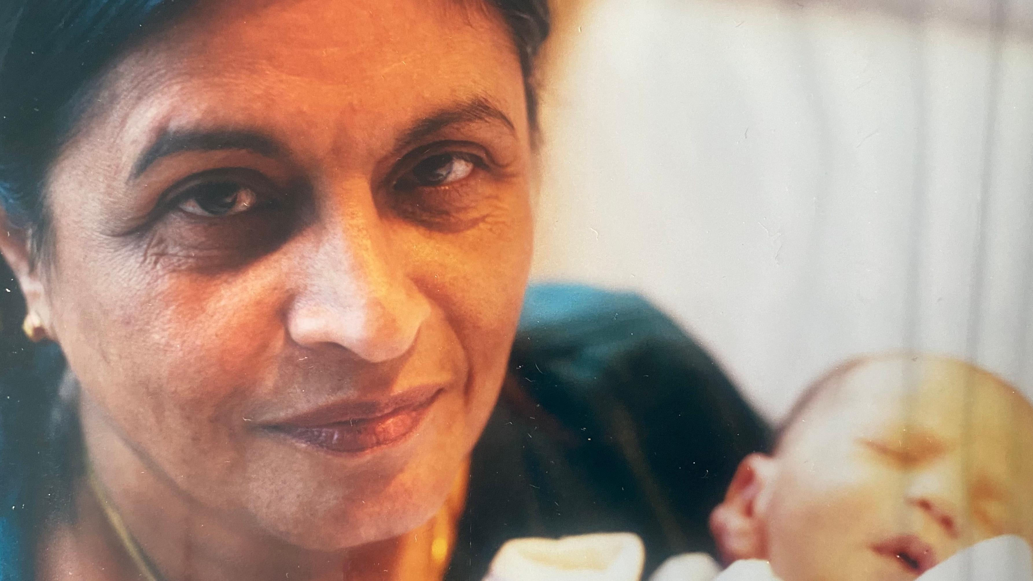 Starija žena gleda ka objektivu i drži bebu u rukama koja spava 