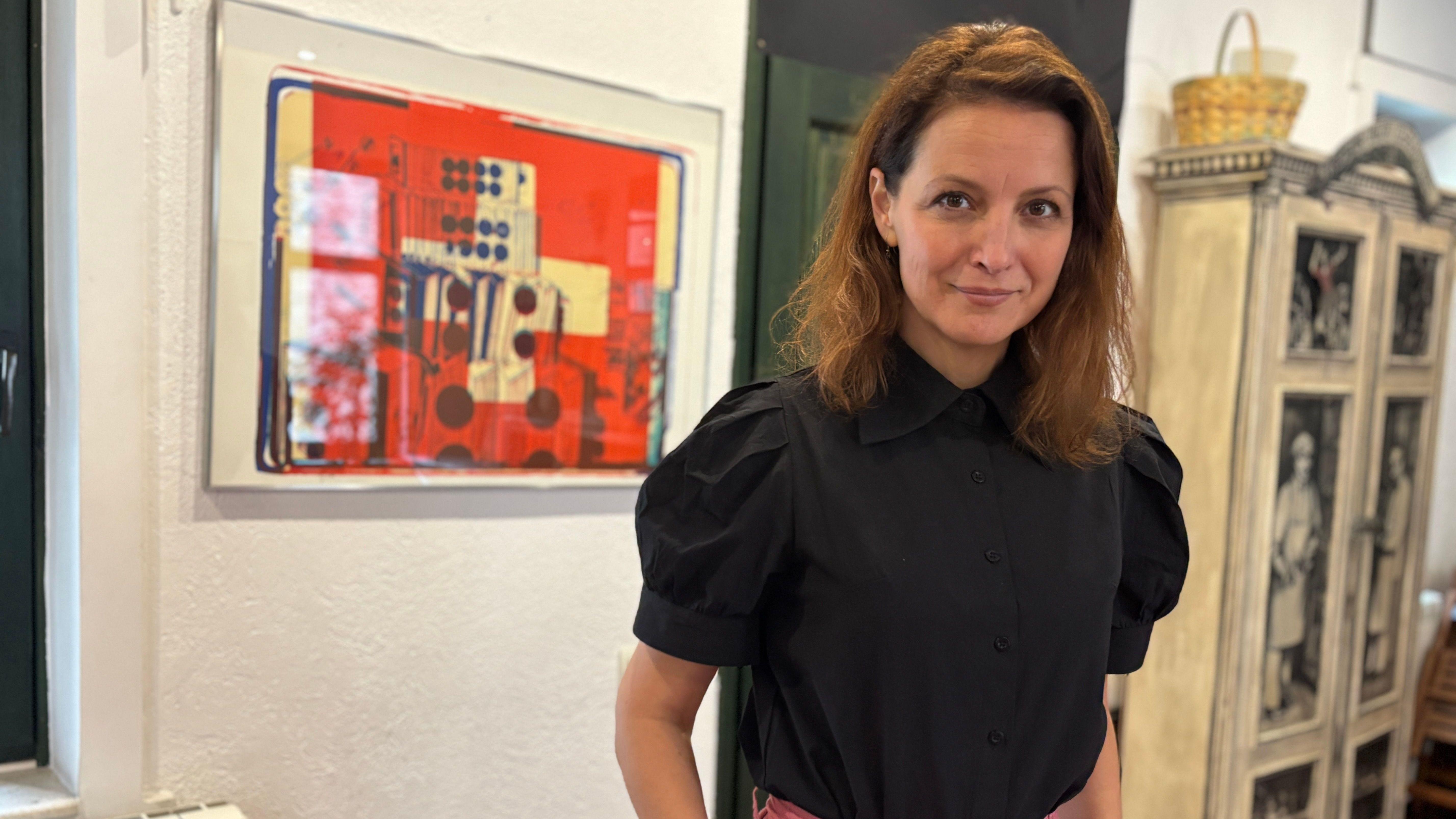Jelena Stupljanin u crnoj košulji i roze pantalonama, sa rukama u džepovima, ima riđu dugu kosu, gleda u kameru i smeška se u kameru, iza nje slika crvene boje i bela vitrina