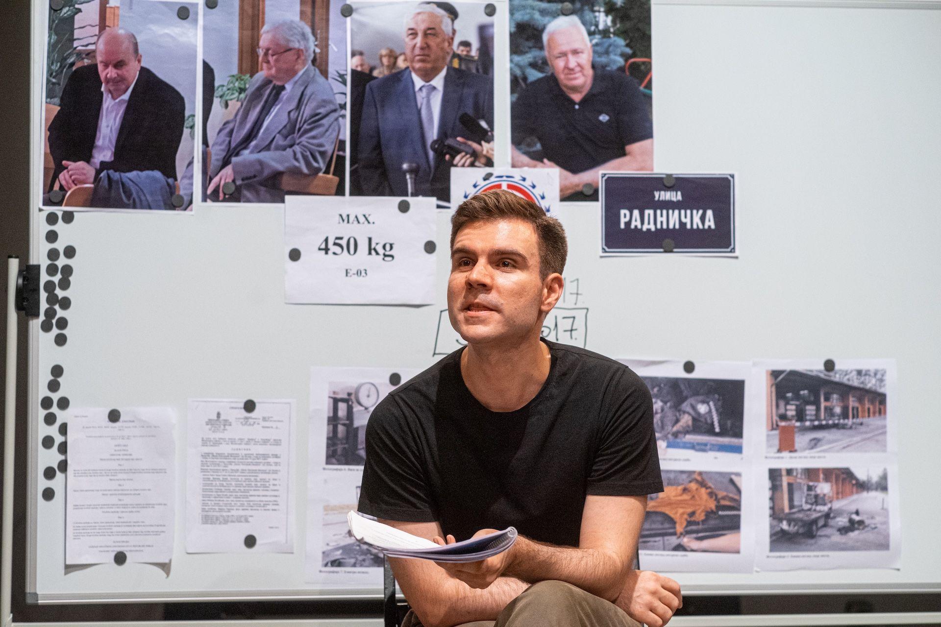 Borisav Matić u crnoj majici, sedi i priča, drži u ruci papire, iza njega pano sa fotografijama i dokumentima
