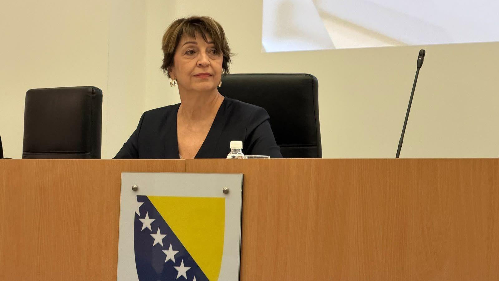 Žena za govornicom, ispred nje mikrofon i grb BiH, 