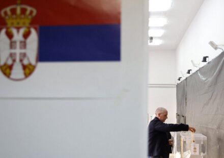 Čovek ubacuje glasački listić u kutiju u krupnom planu zastava Srbije