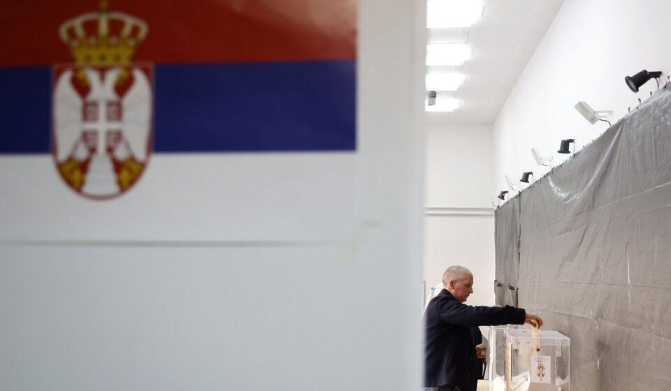 Čovek ubacuje glasački listić u kutiju u krupnom planu zastava Srbije