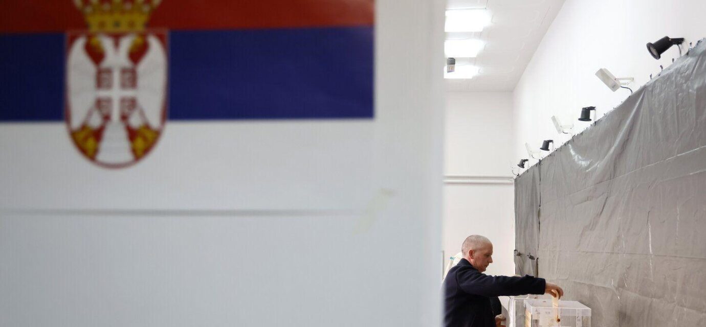 Čovek ubacuje glasački listić u kutiju u krupnom planu zastava Srbije
