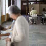Dvoje ljudi sedi iza kartonskog paravana, starija žena i dvoje mladih sa papirima
