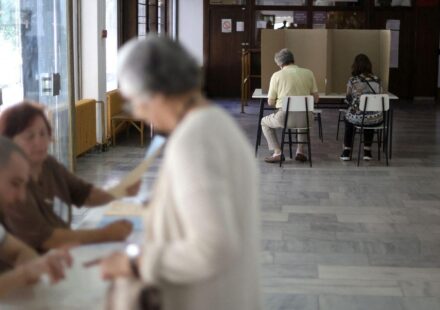 Dvoje ljudi sedi iza kartonskog paravana, starija žena i dvoje mladih sa papirima