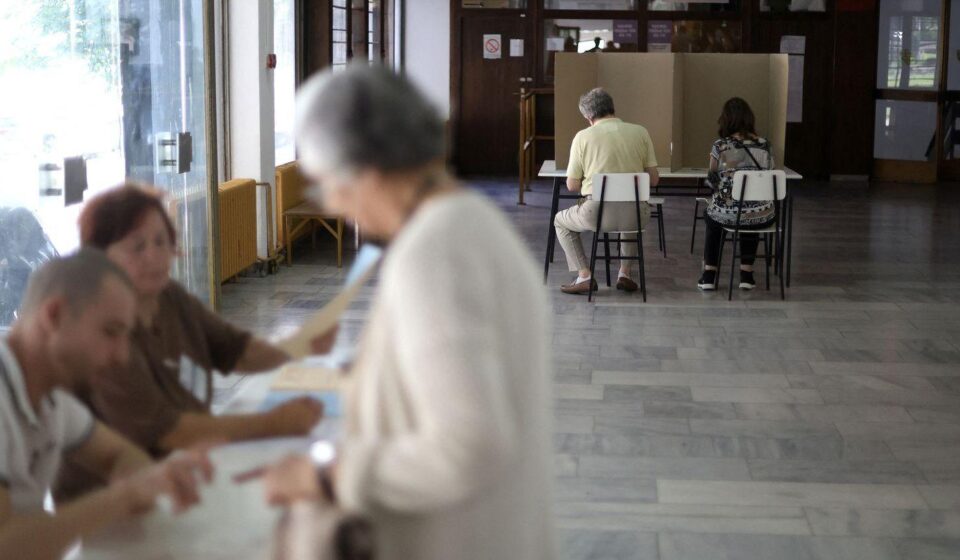 Dvoje ljudi sedi iza kartonskog paravana, starija žena i dvoje mladih sa papirima