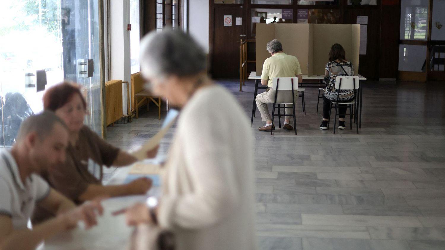 Dvoje ljudi sedi iza kartonskog paravana, starija žena i dvoje mladih sa papirima