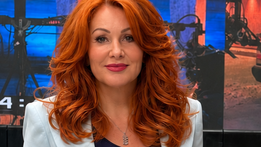 Žena sa narandžastom kosom u odelu, našminkana, gleda u kameru, ima oko vrata srebrni lančić