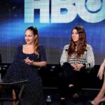 Four actresses sit in a line on black chairs while on stage with the lettering HBO seen in the background.
