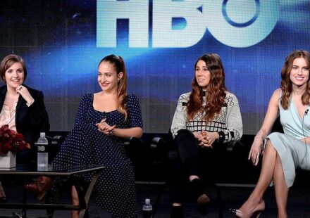 Four actresses sit in a line on black chairs while on stage with the lettering HBO seen in the background.