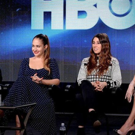 Four actresses sit in a line on black chairs while on stage with the lettering HBO seen in the background.