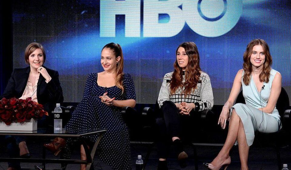 Four actresses sit in a line on black chairs while on stage with the lettering HBO seen in the background.