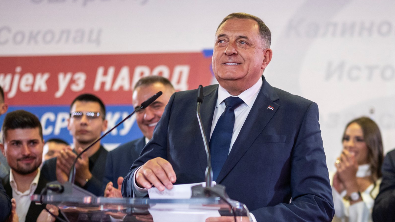 Milorad Dodik u plavom odelu i sa osmehom na usnama za govornicom, drži papire u rukama